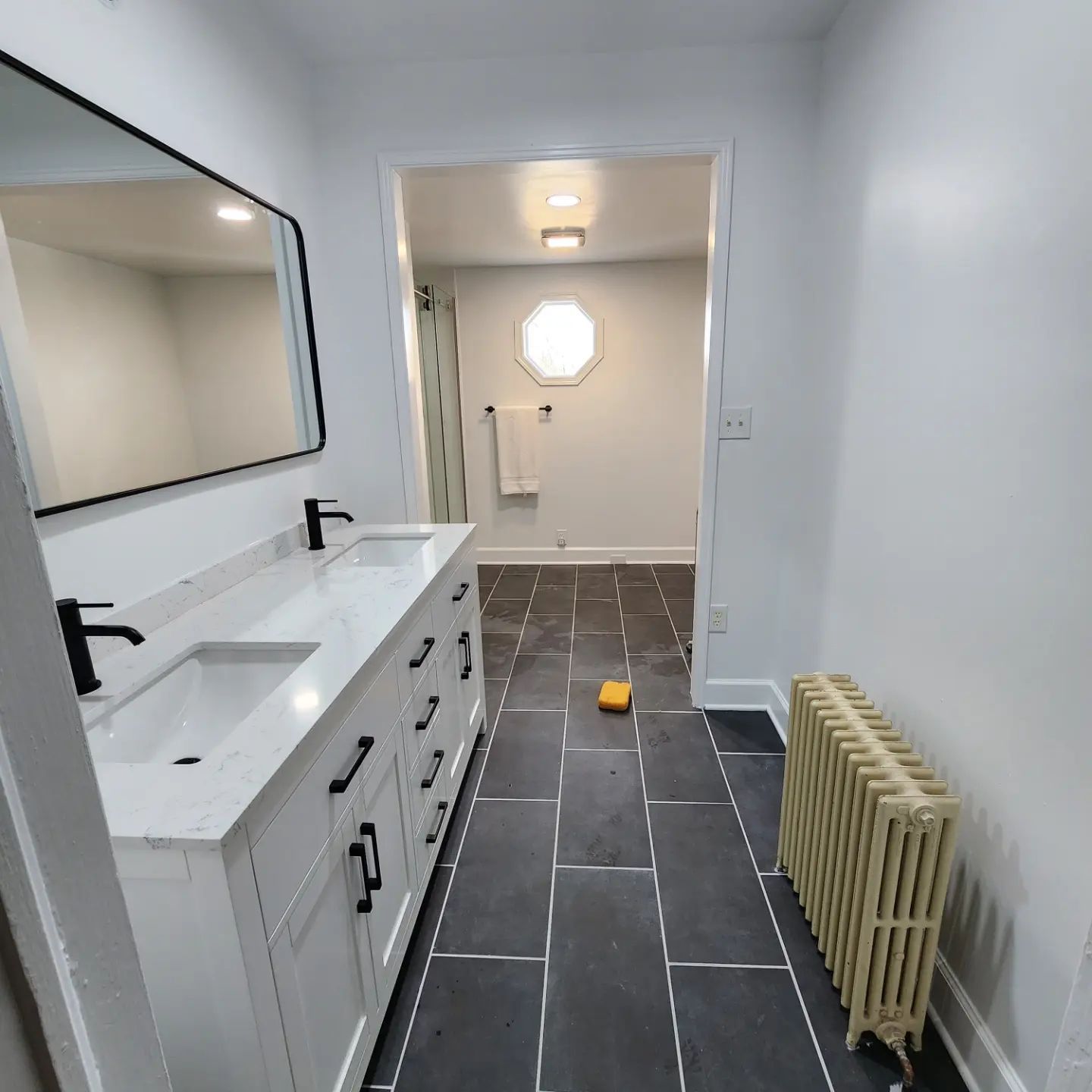 Modern bathroom with double vanity, dark tile floor, and a yellow bucket in the hallway beyond.