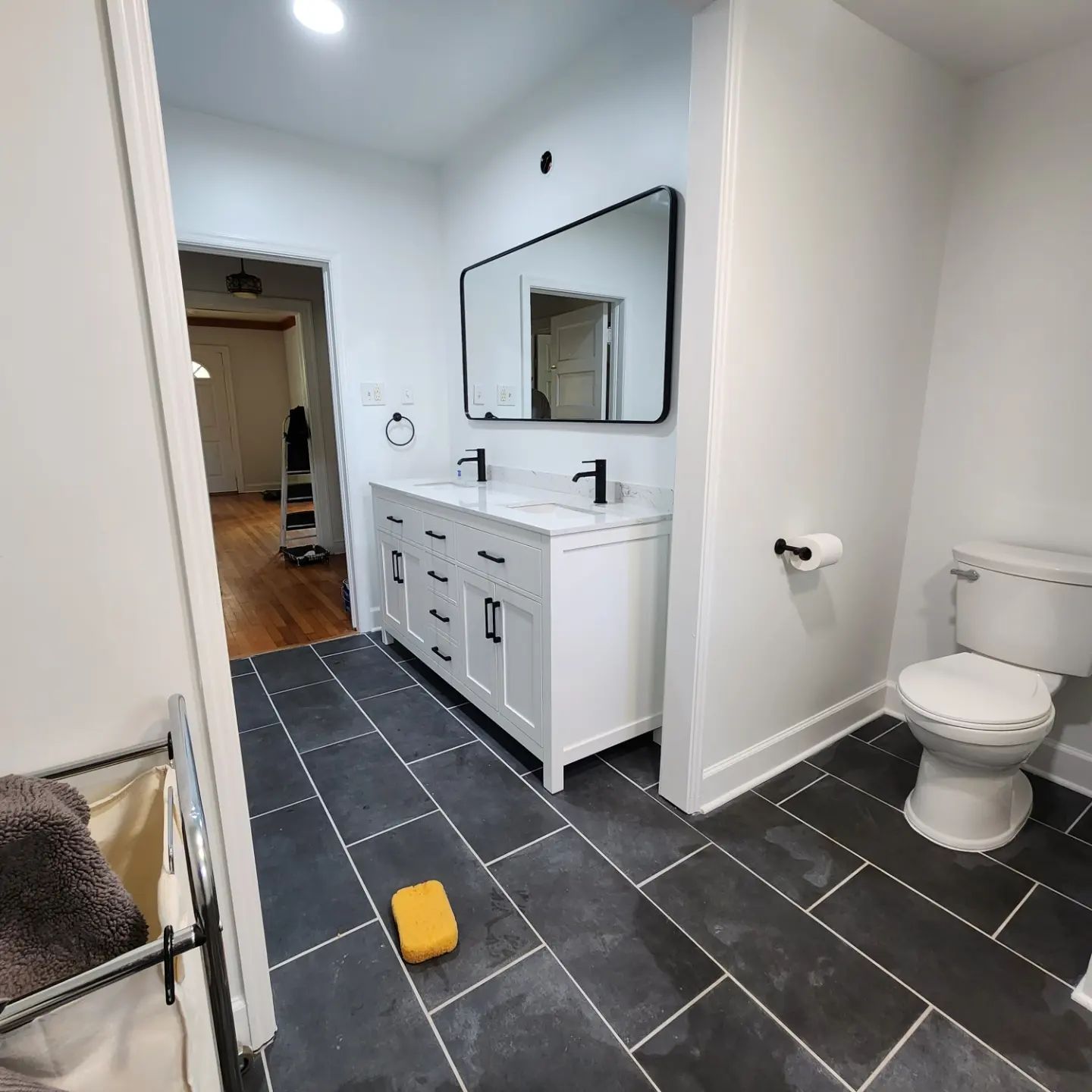 Modern bathroom with black tile floor, white vanity, mirror, toilet, and a yellow sponge on the floor