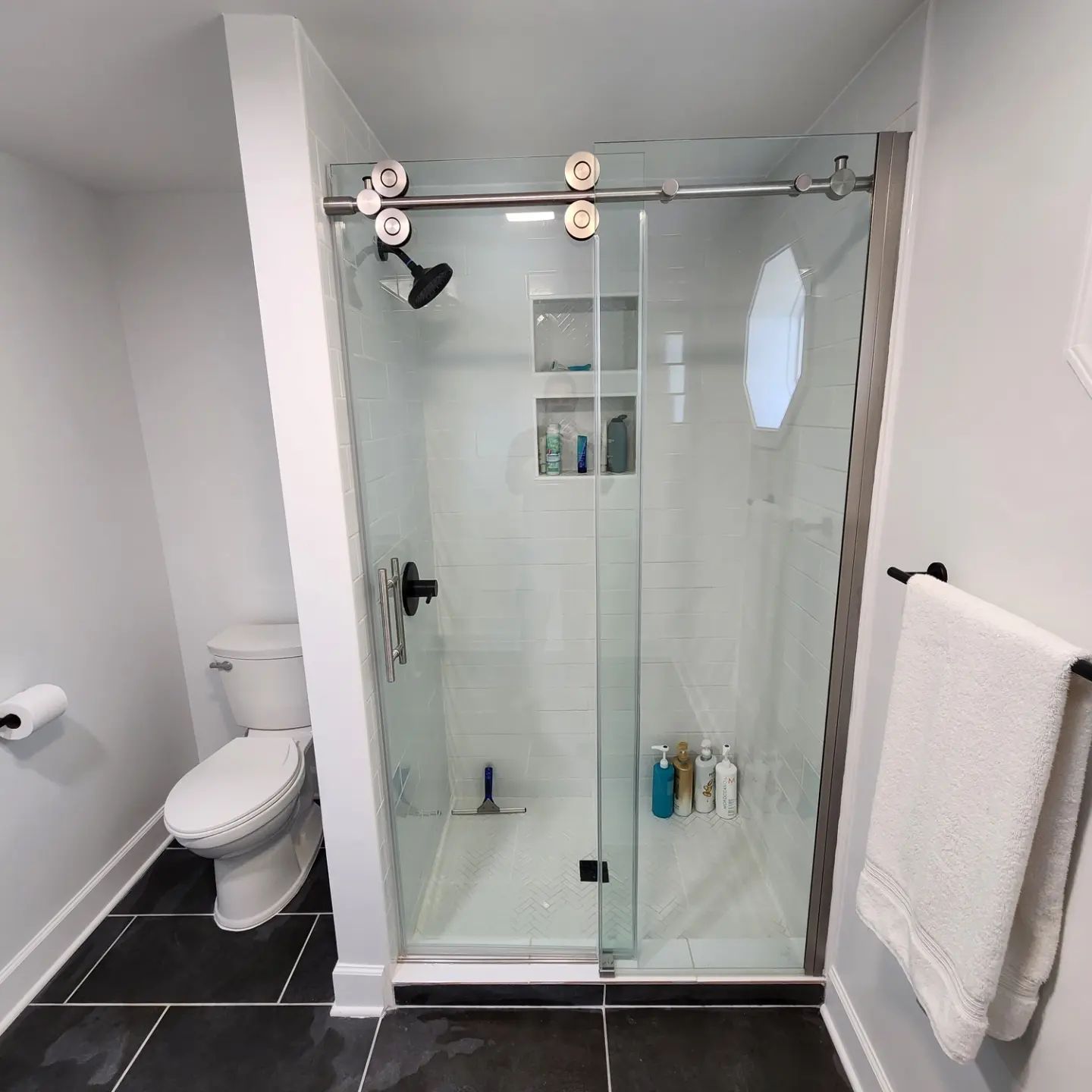Compact modern bathroom with glass shower, toilet, black tile floor, and white towels on a wall rack