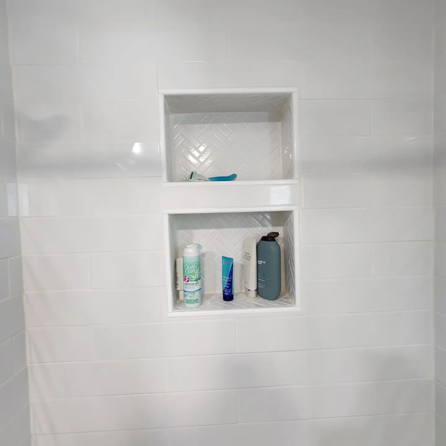 White tiled shower wall with two recessed shelves holding toiletries and a blue loofah.
