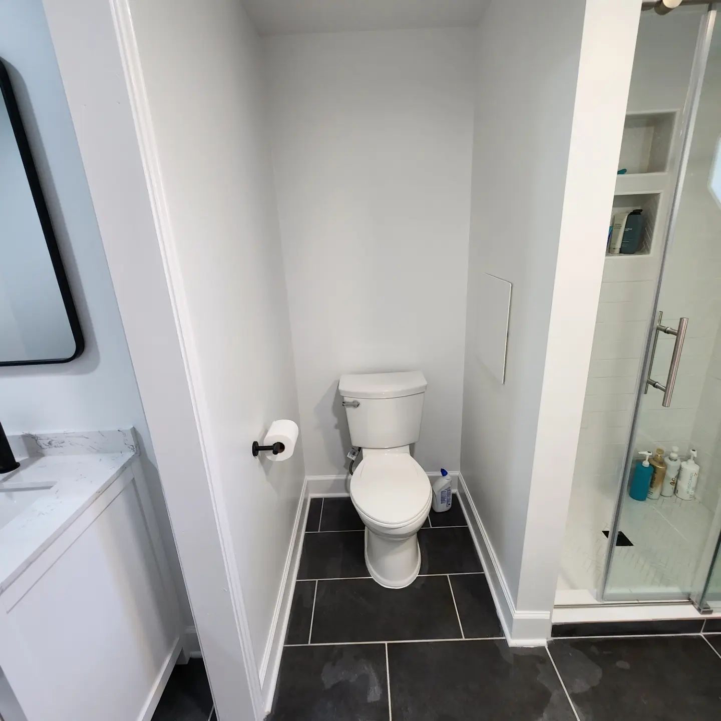 Small white bathroom with a toilet, black tile floor, and glass shower on the right.