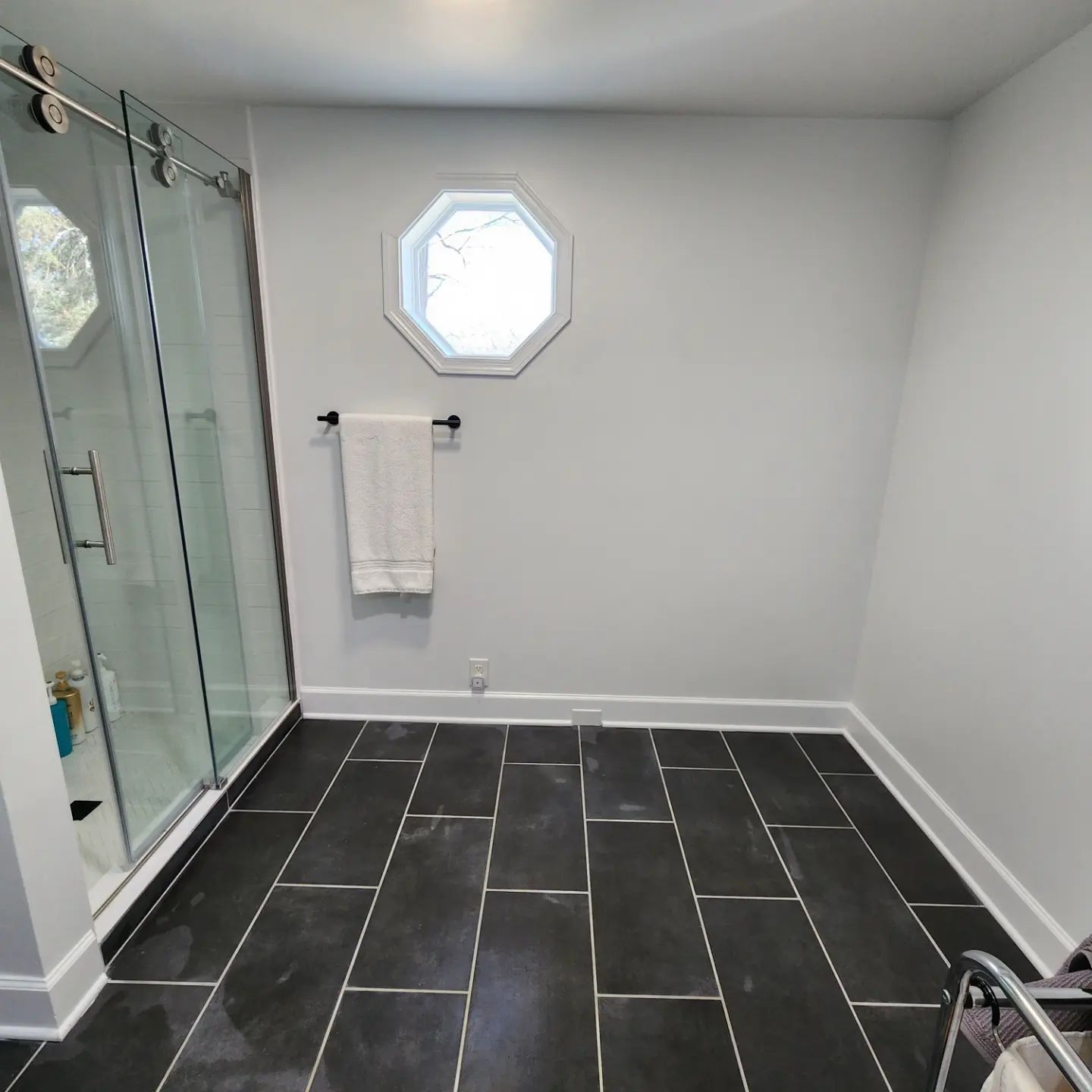 Modern bathroom with dark tile floor, glass shower, towel rack, and octagonal window