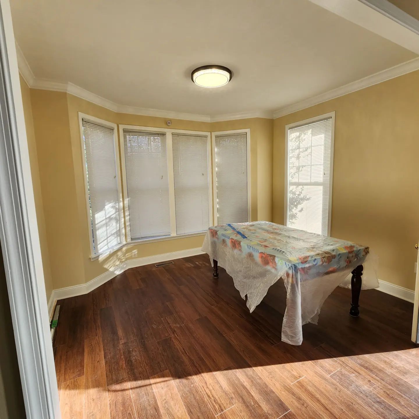 Empty dining room with bay windows, beige walls, hardwood floor, and a table covered in a floral cloth.
