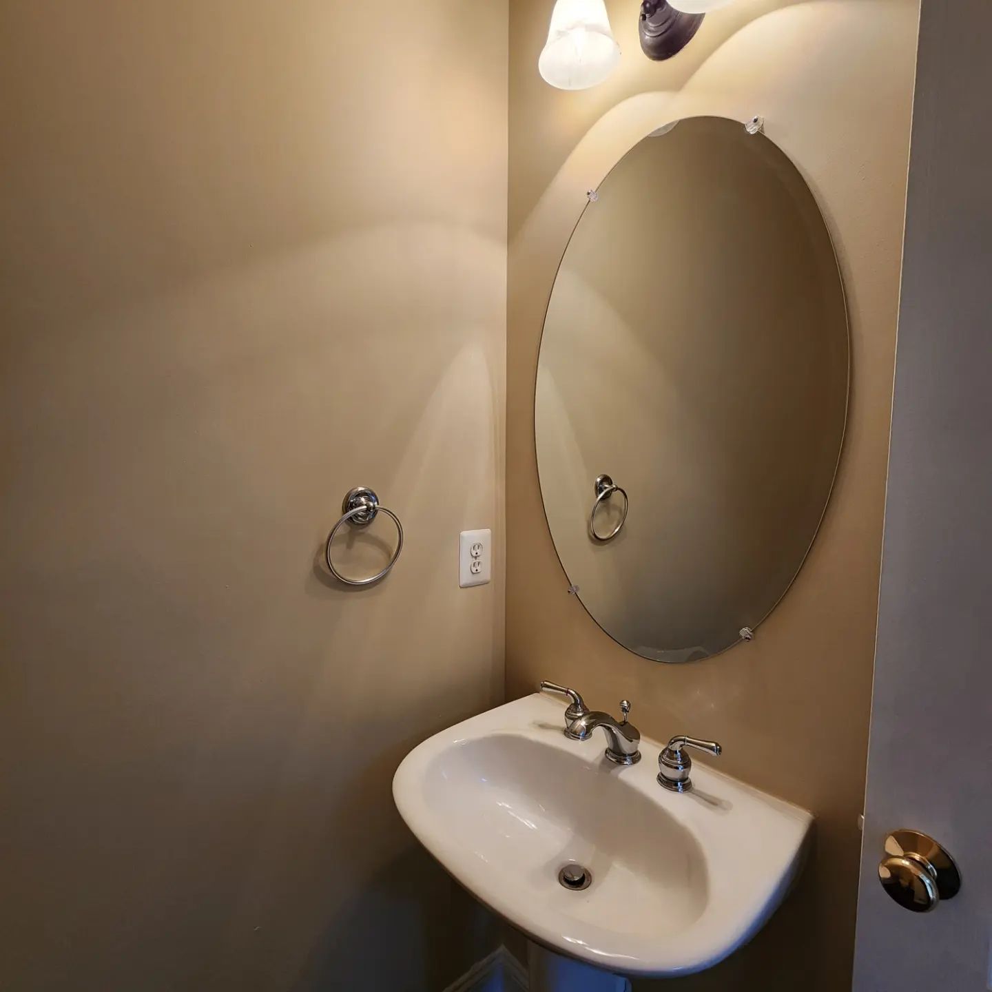 Small beige bathroom with oval mirror, sink, towel ring, and light fixture.