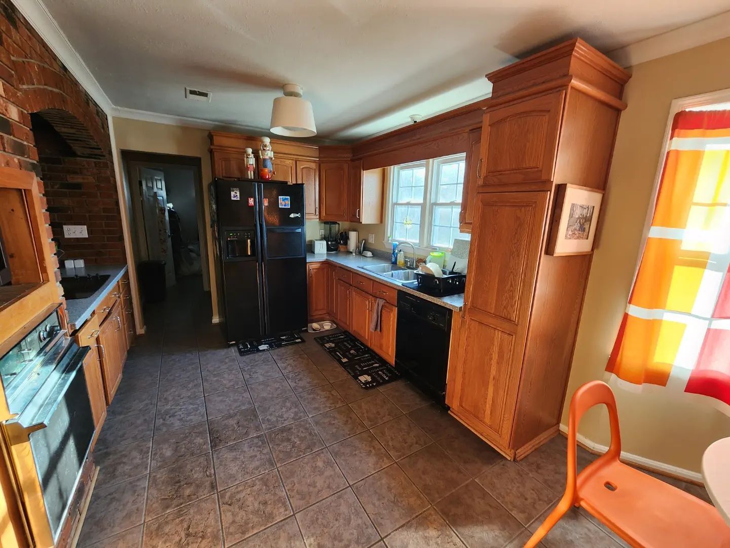 Narrow kitchen with wood cabinets, black fridge, brick wall, tiled floor, and orange chair by bright window.