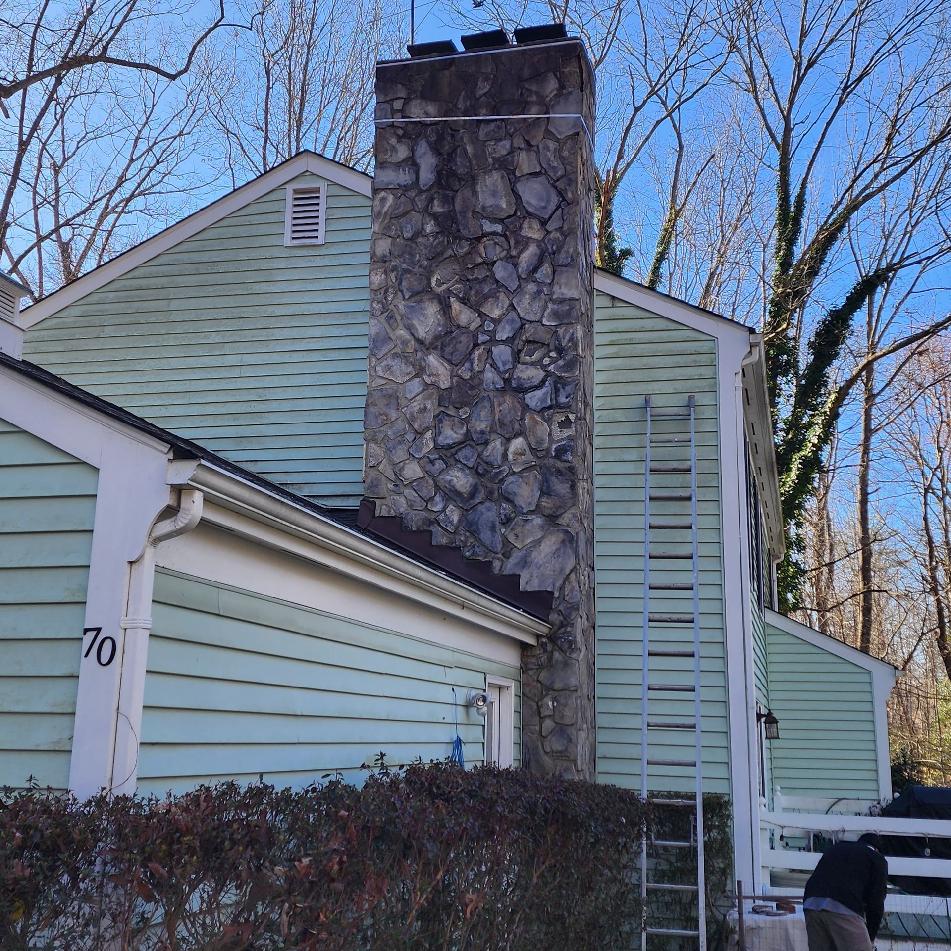 Light green house with a tall stone chimney and leafless trees under a clear blue sky
