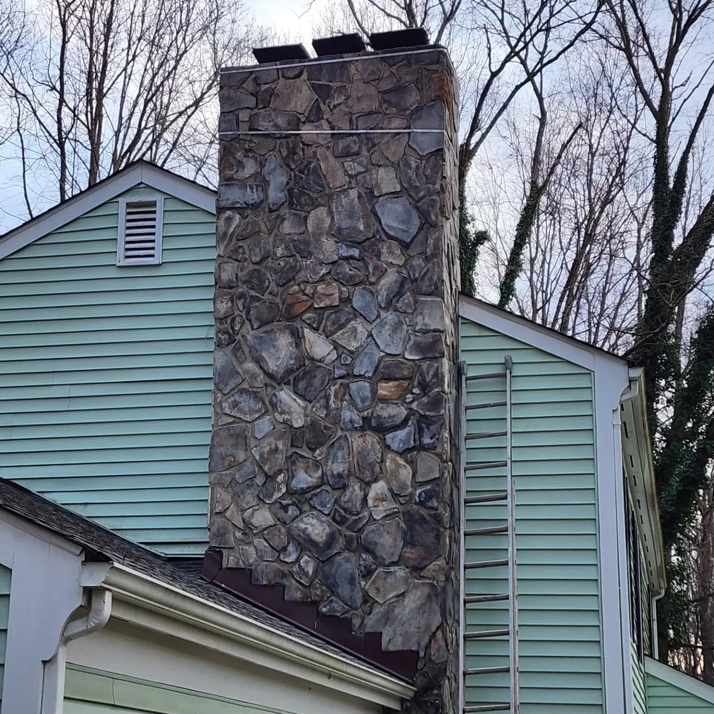 Stone chimney on a light green house with leafless trees in the background.
