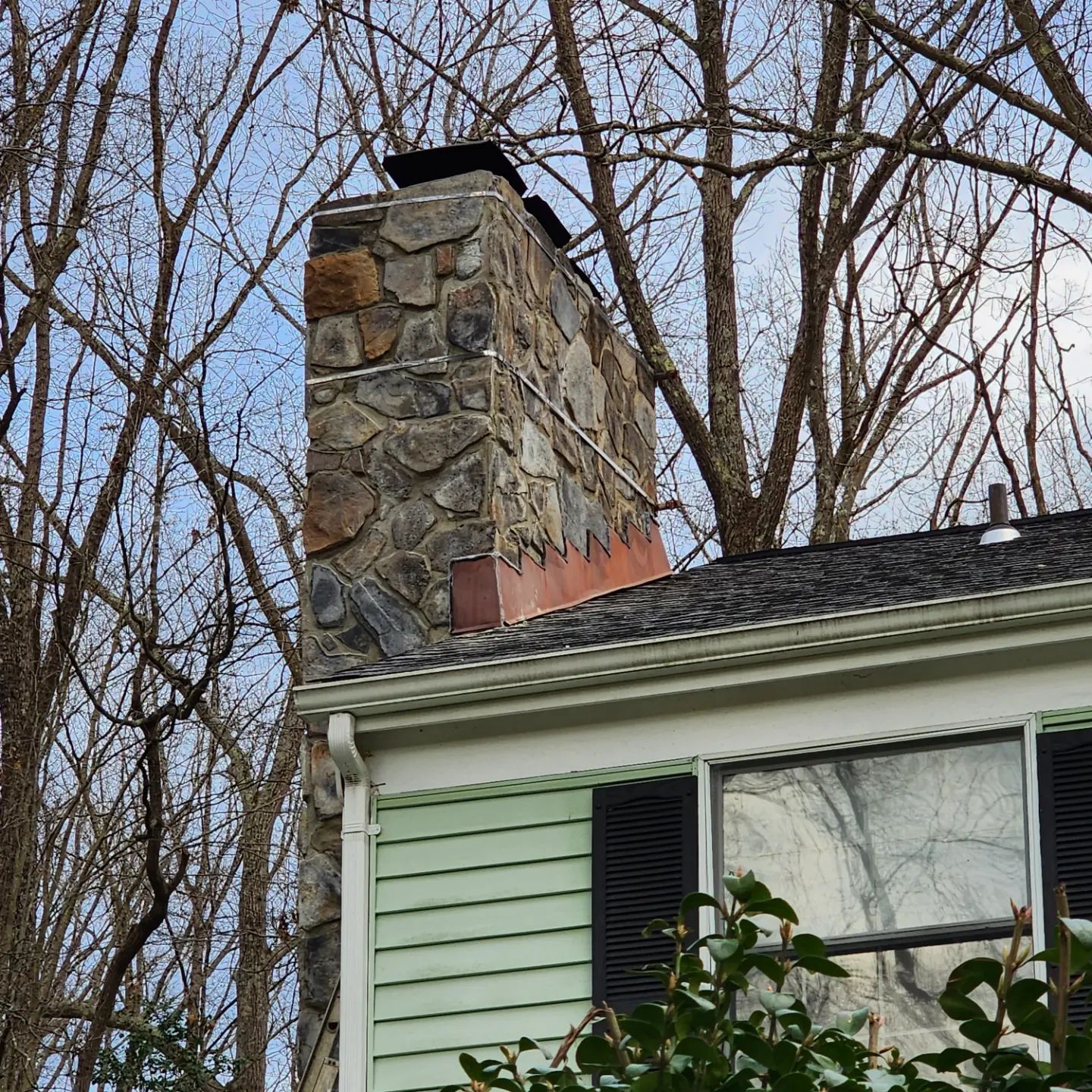 Stone chimney on a pale green house with black shutters, seen against bare winter trees.