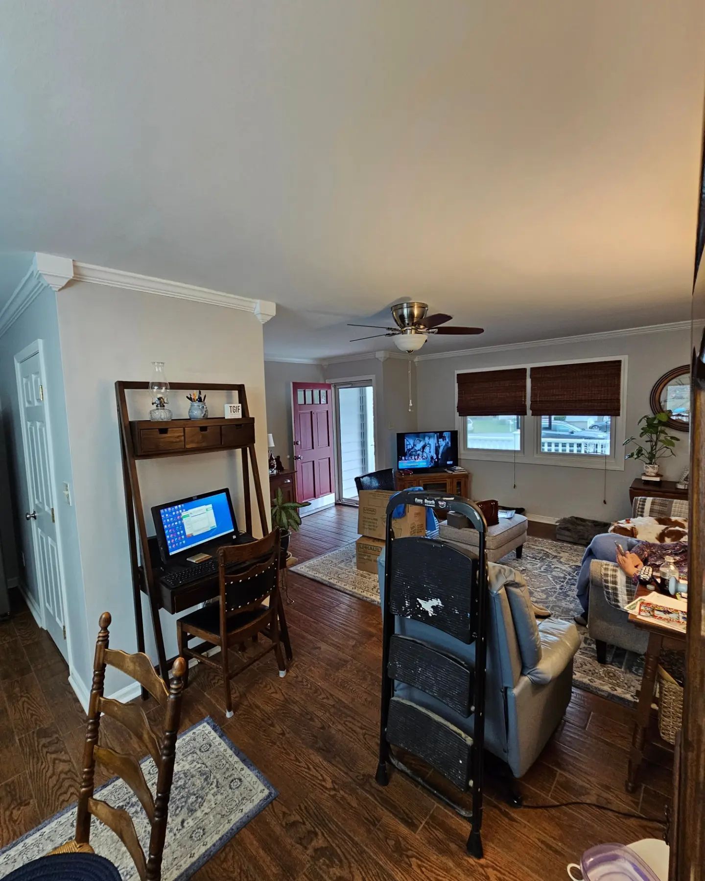 Cluttered living room with TV, desk, chairs, hardwood floors, and windows with blinds and curtains