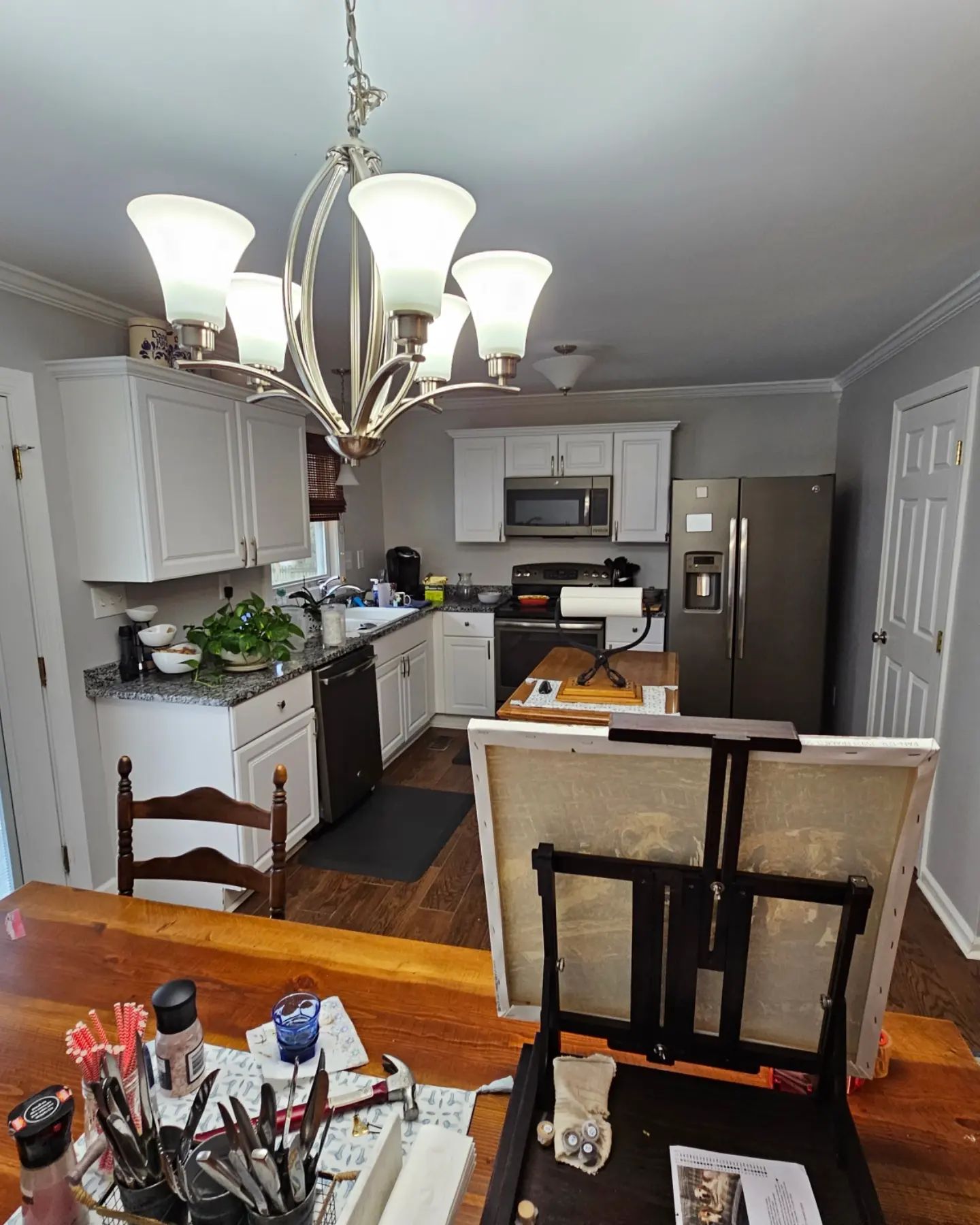 Kitchen seen from dining area, with chandelier, white cabinets, stainless fridge, and a cluttered table in foreground