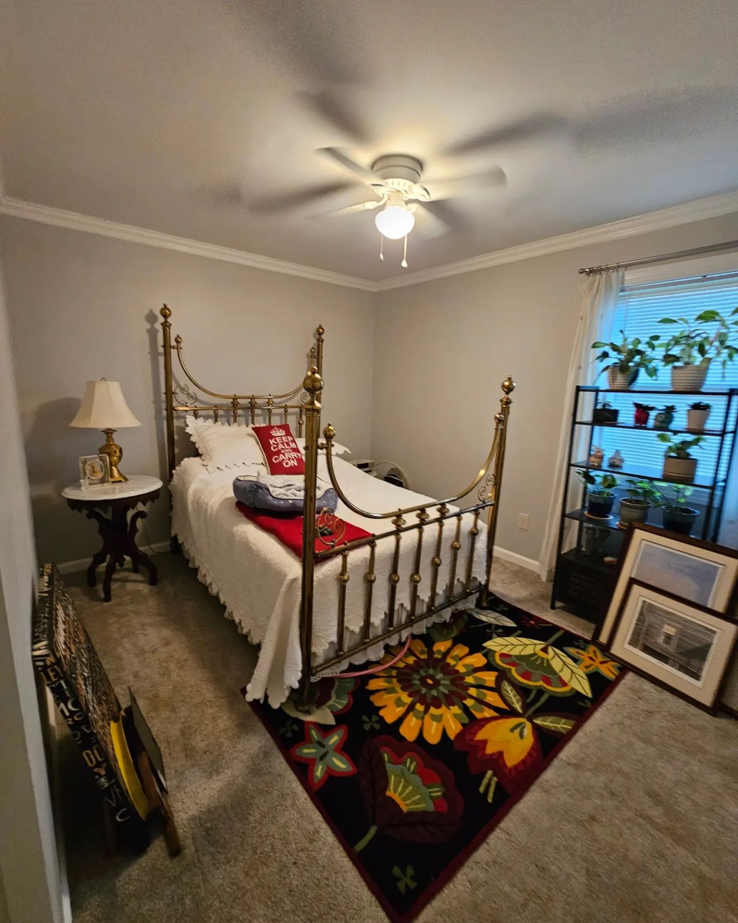 Vintage brass bed in a cozy bedroom with colorful quilt, ceiling fan, lamp, and window plants.