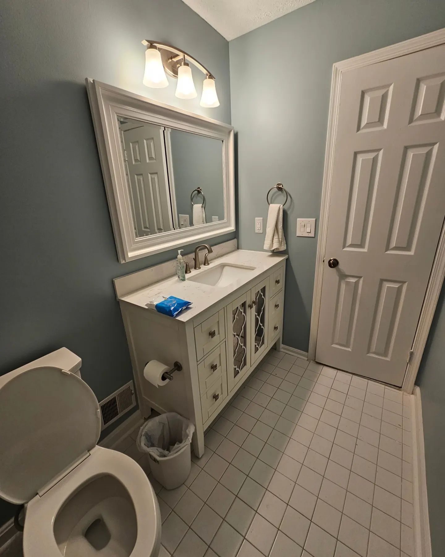 Small bathroom with white vanity, mirror lights, toilet, towel ring, and gray-blue walls.