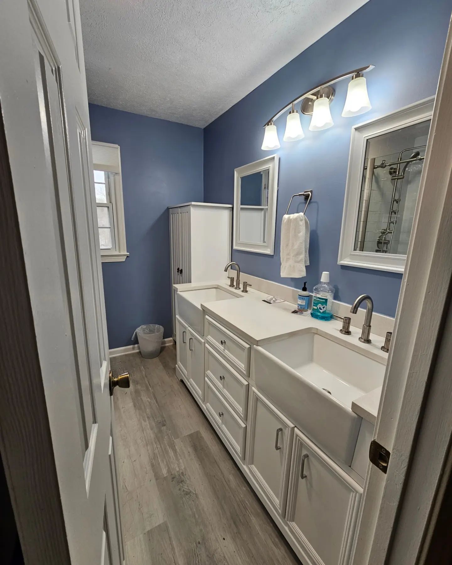 Narrow blue bathroom with white vanity, sink, mirror, towel, and window on the left.