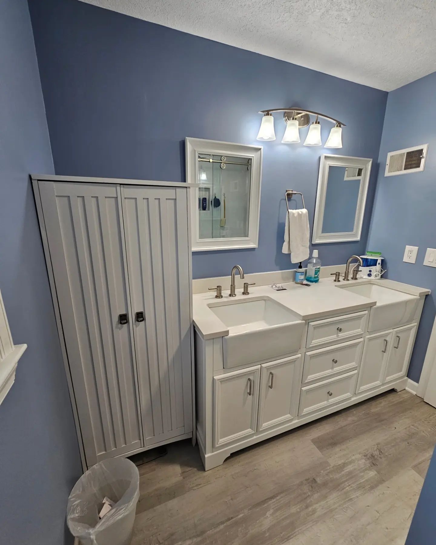 Blue bathroom with double vanity, white cabinets, mirrors, and a towel rack beside a tall storage cabinet.