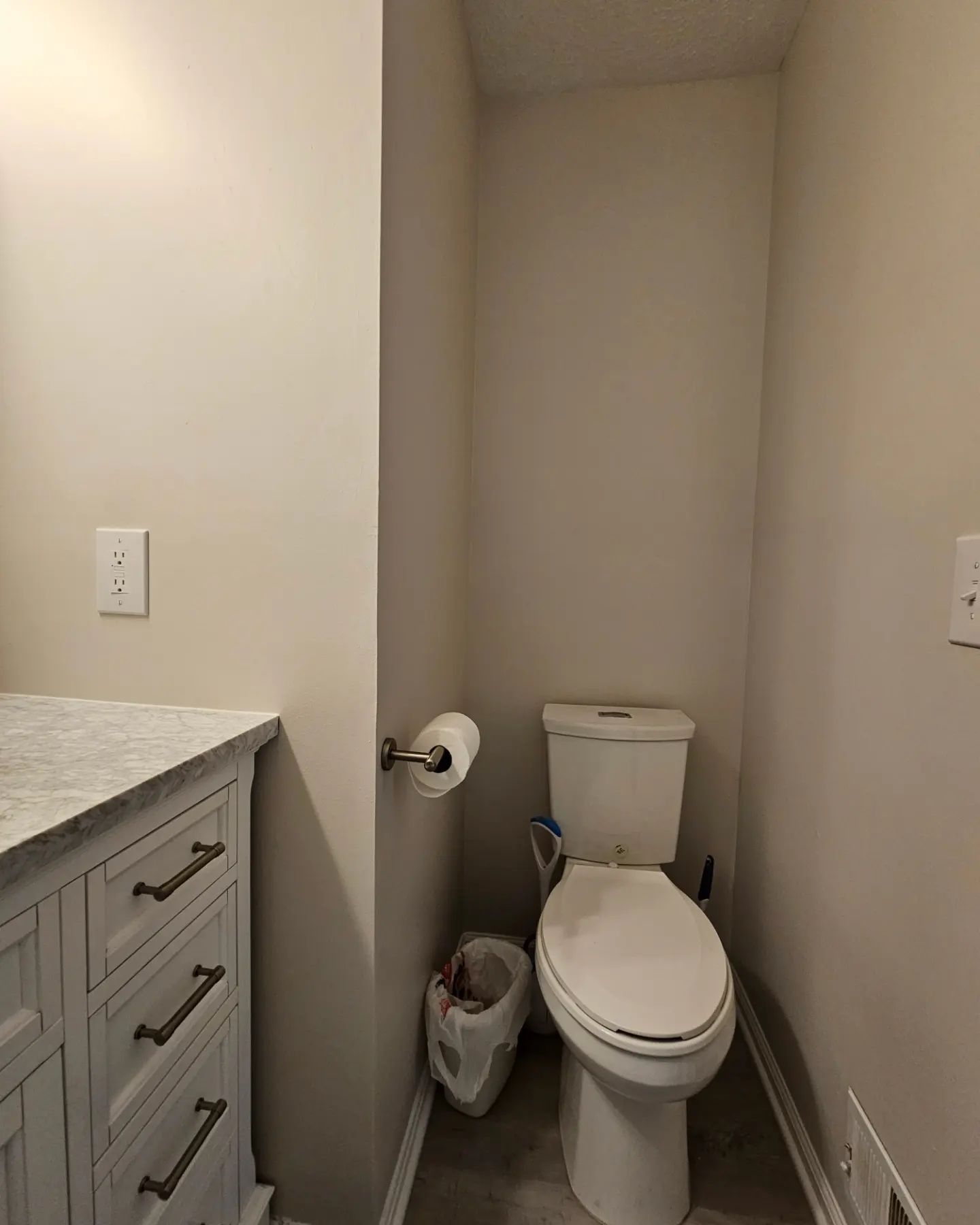 Small narrow bathroom with a toilet, trash can, and gray vanity beside a beige wall.