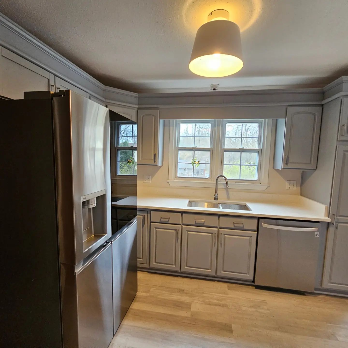 Small gray kitchen with white countertops, stainless fridge, sink under windows, and wood floor.