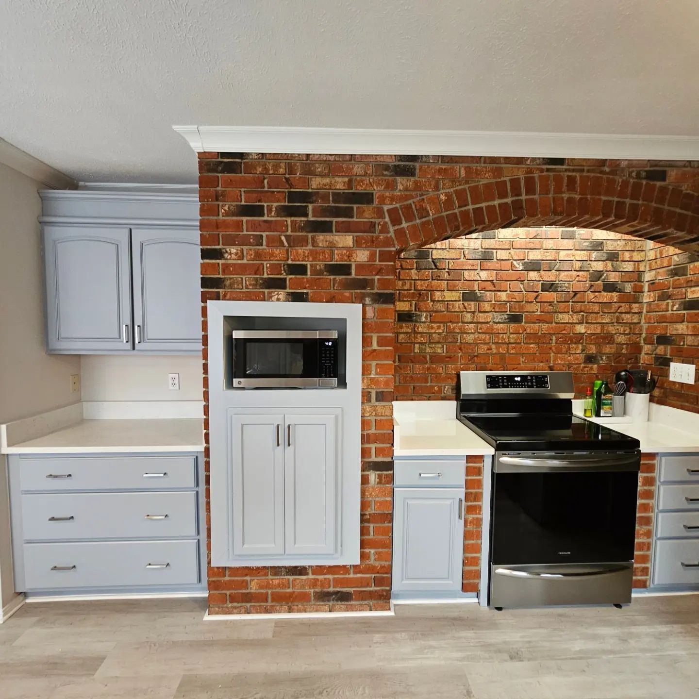 Kitchen with brick arch wall, gray cabinets, white counters, built-in microwave, and black stove on the right.