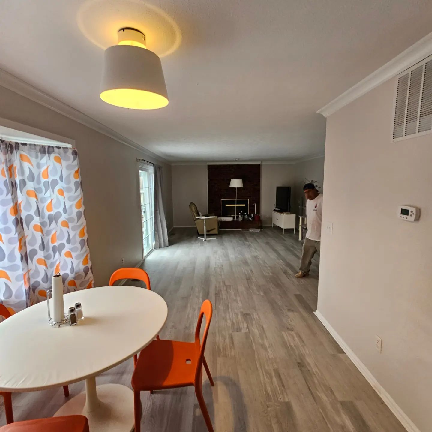 Bright living-dining room with orange chairs, round table, gray wood floors, and a person standing near the back wall