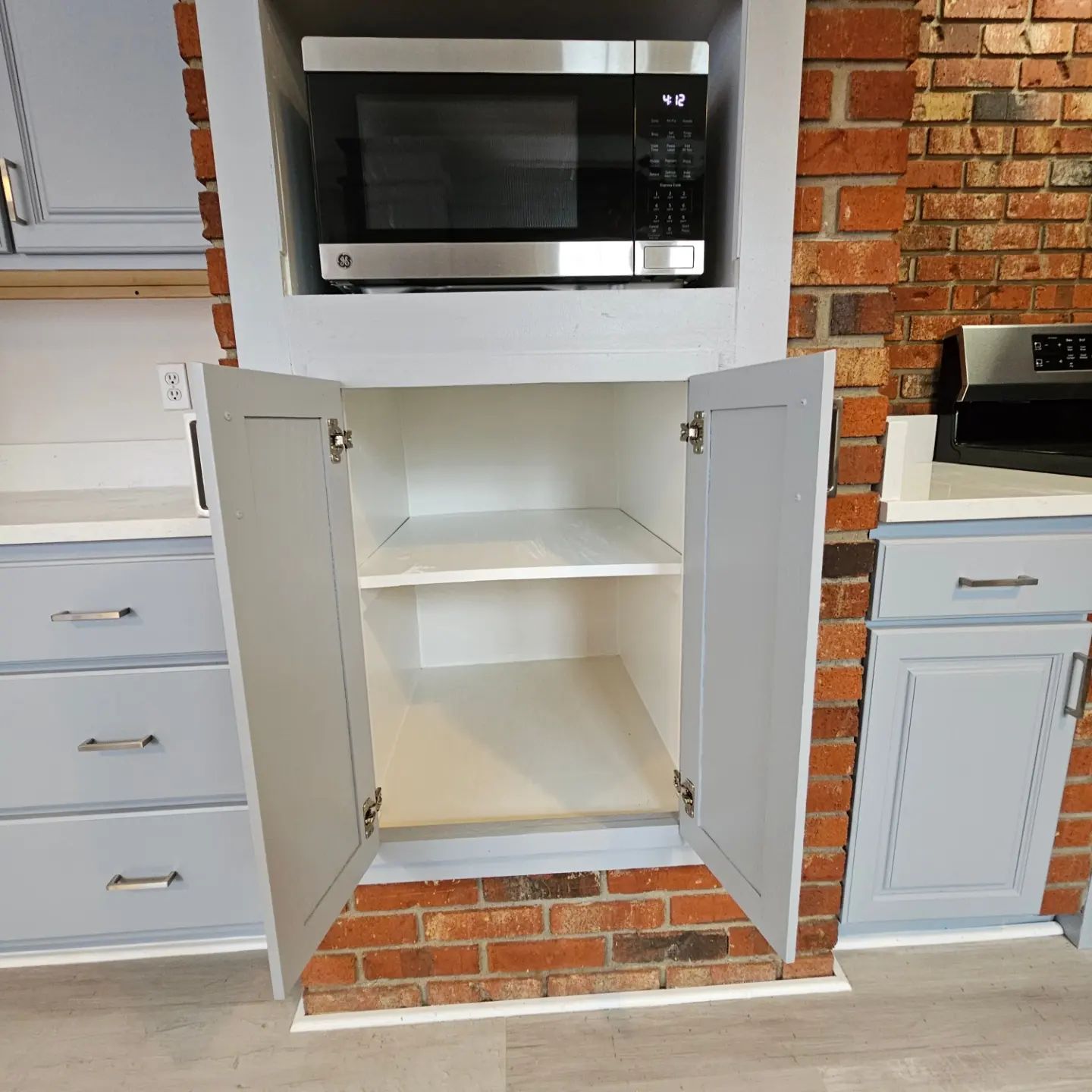 Open kitchen cabinet with shelves beneath a built-in microwave, framed by white cabinets and brick trim.
