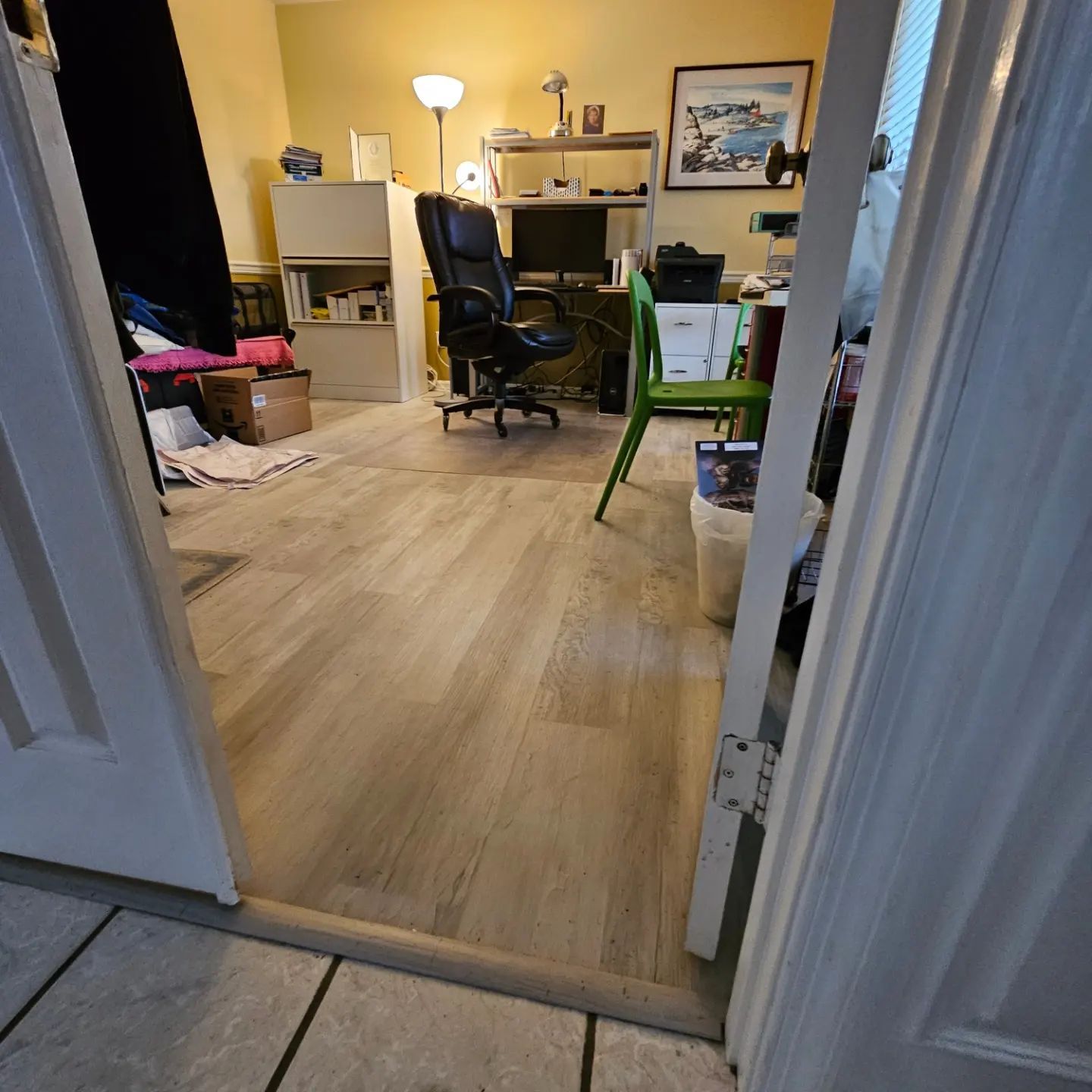 Open doorway into a cluttered home office with a desk, chair, lamp, and green step stool on wood flooring.