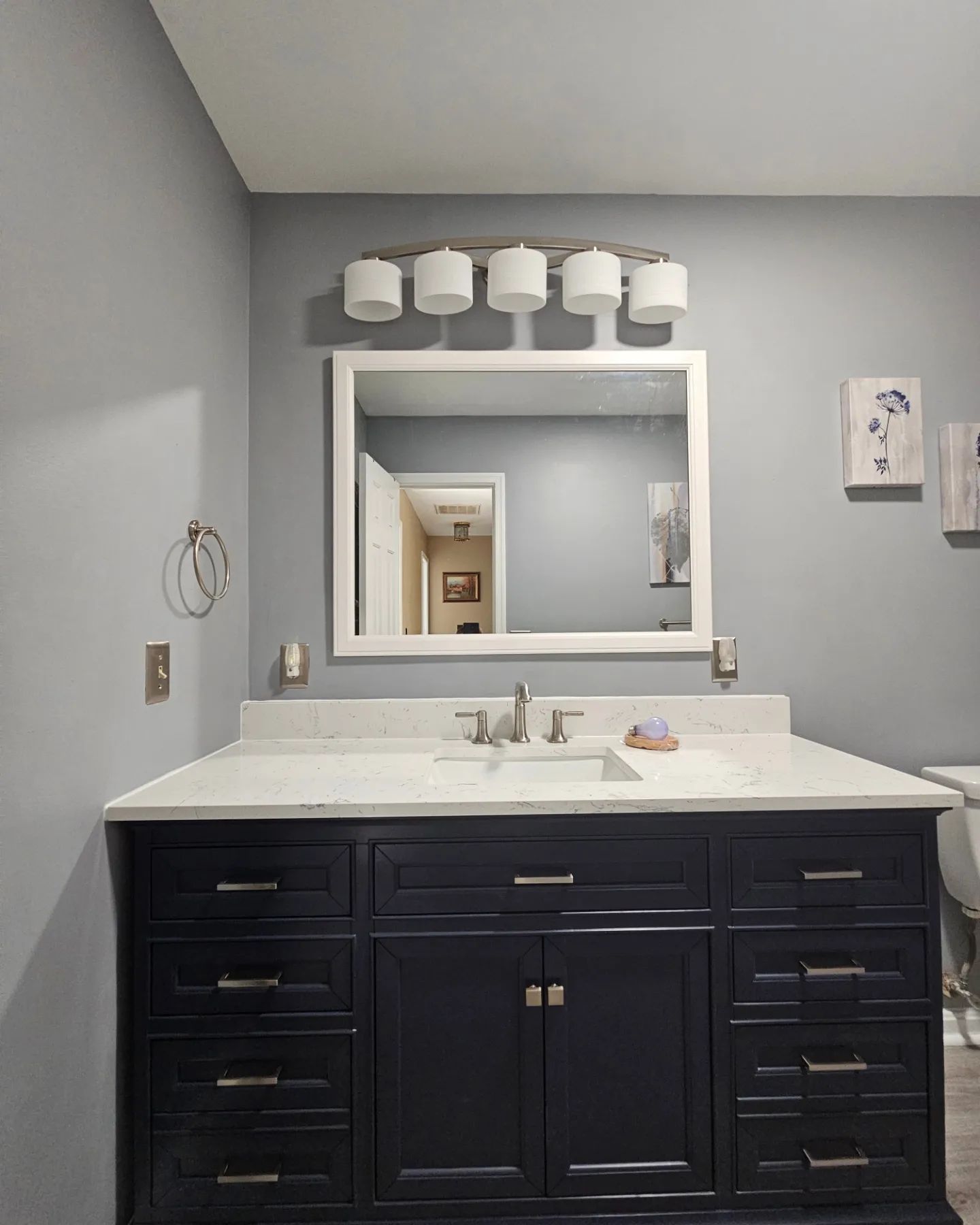 Bathroom vanity with white countertop, dark cabinets, mirror, and wall lights in a gray room