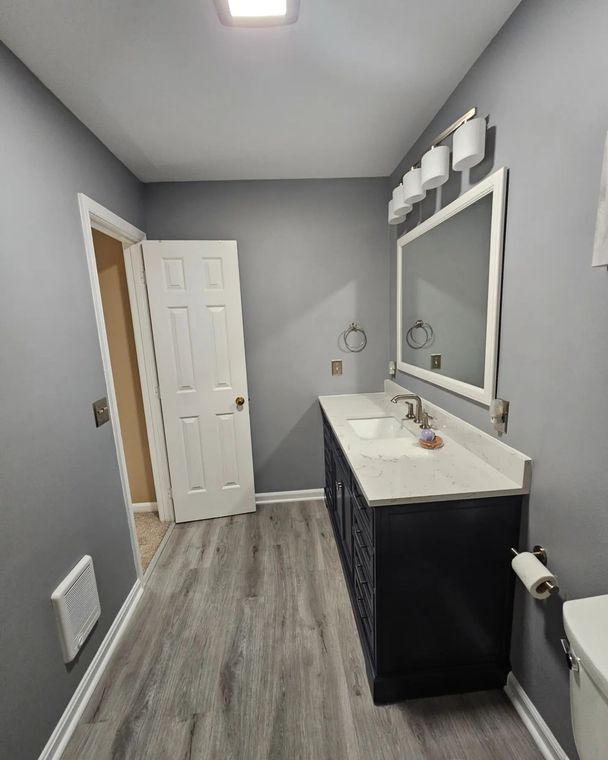 Small gray bathroom with white vanity, mirror, toilet, and wood-look flooring.