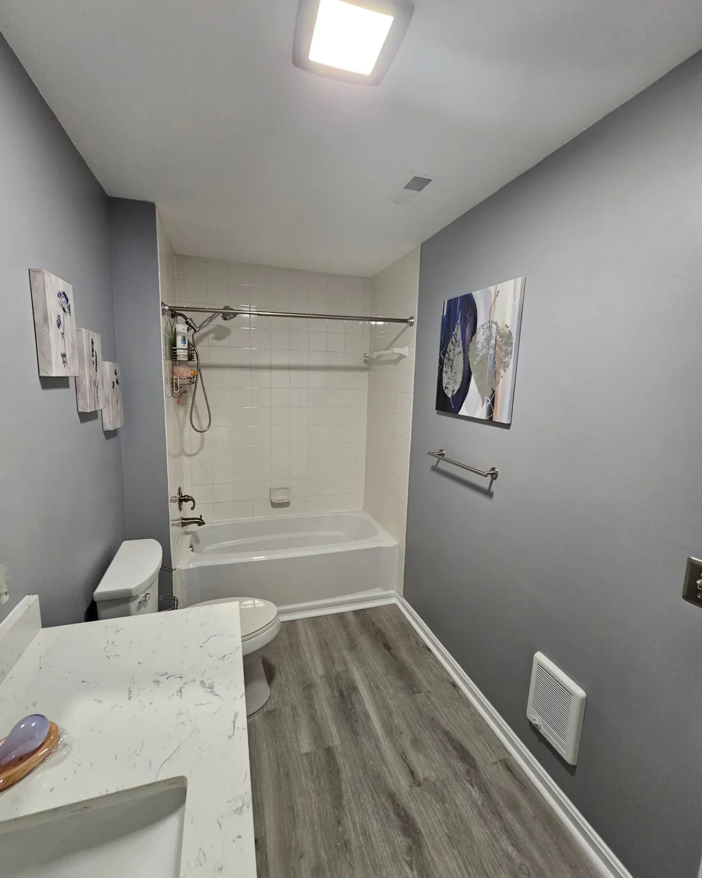 Small bathroom with gray walls, white tub, sink vanity, wood-look floor, and wall art.