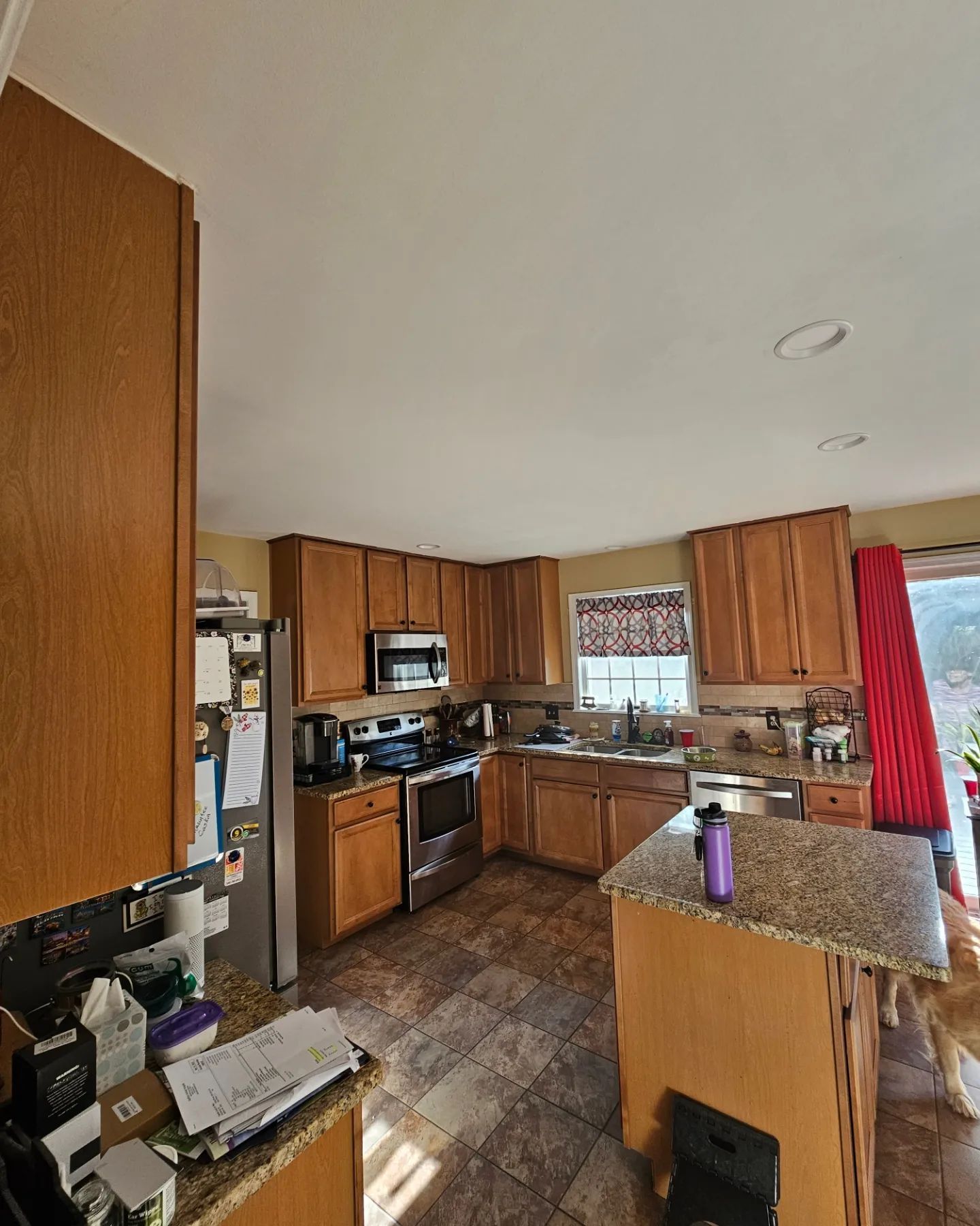 Small kitchen with wood cabinets, granite island, stove, sink, and red curtains.