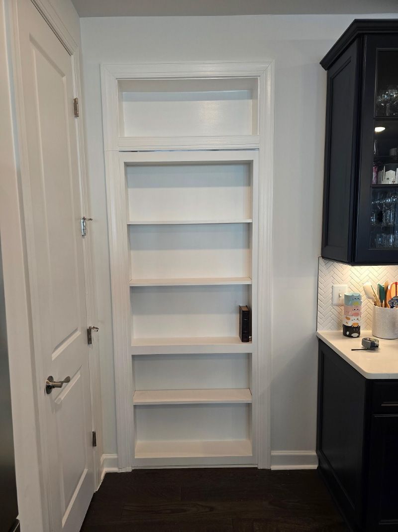 Narrow white built-in shelves in a hallway, flanked by a door and dark kitchen cabinets.