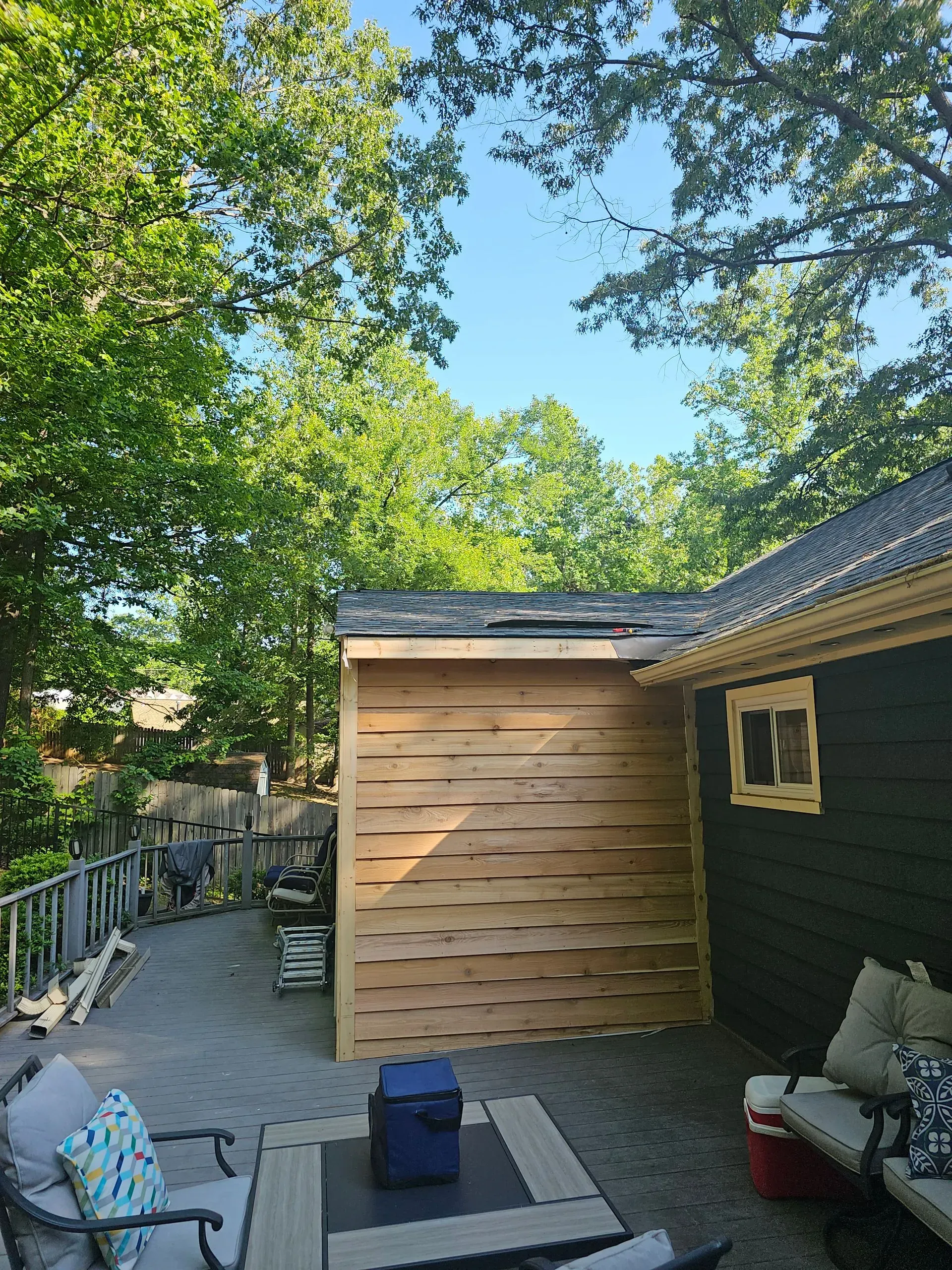 Wooden shed extension beside a house, with a narrow walkway, trees overhead, and a blue bin in the foreground.