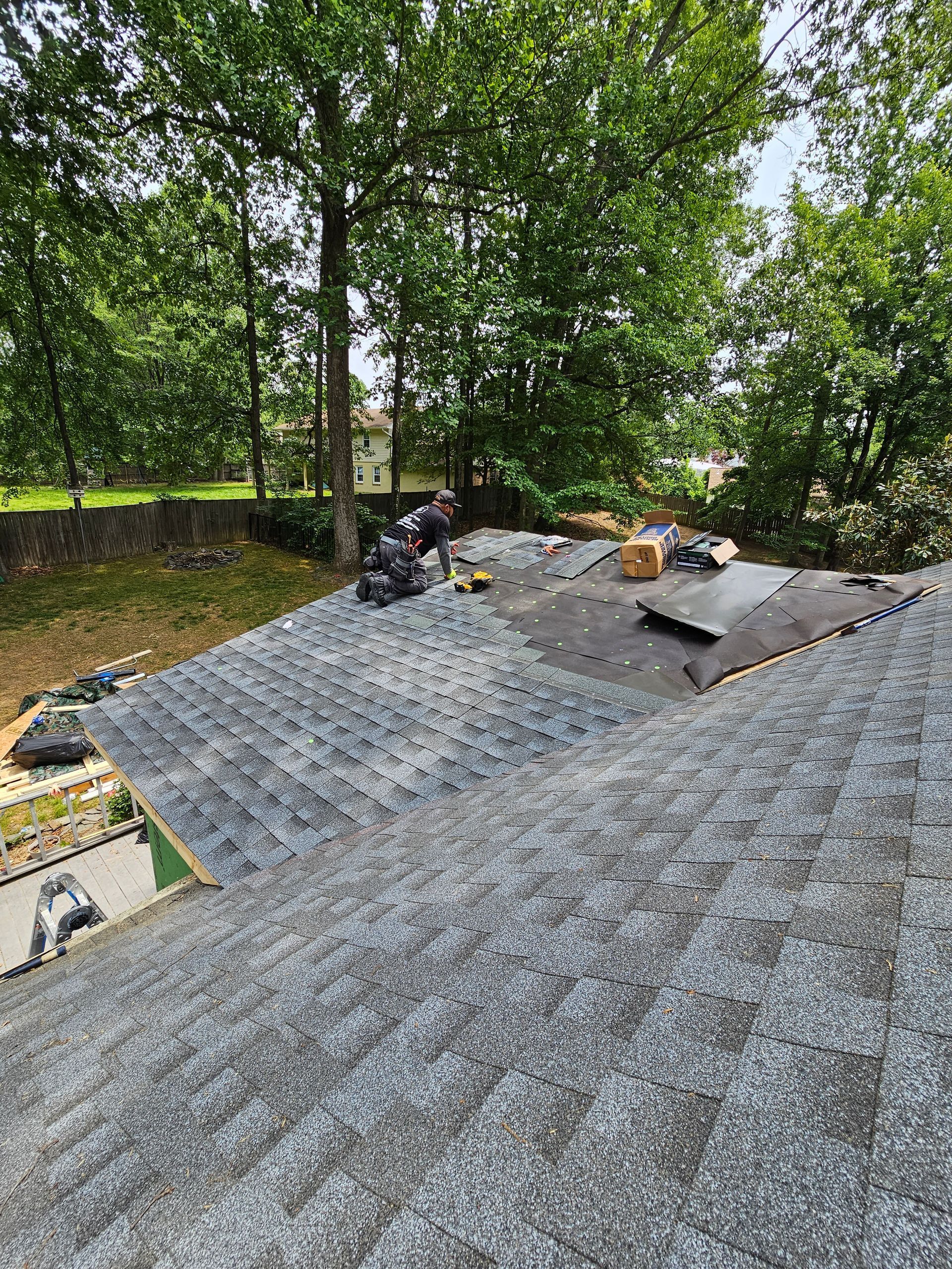 Roof under construction with gray shingles and workers’ materials beside trees