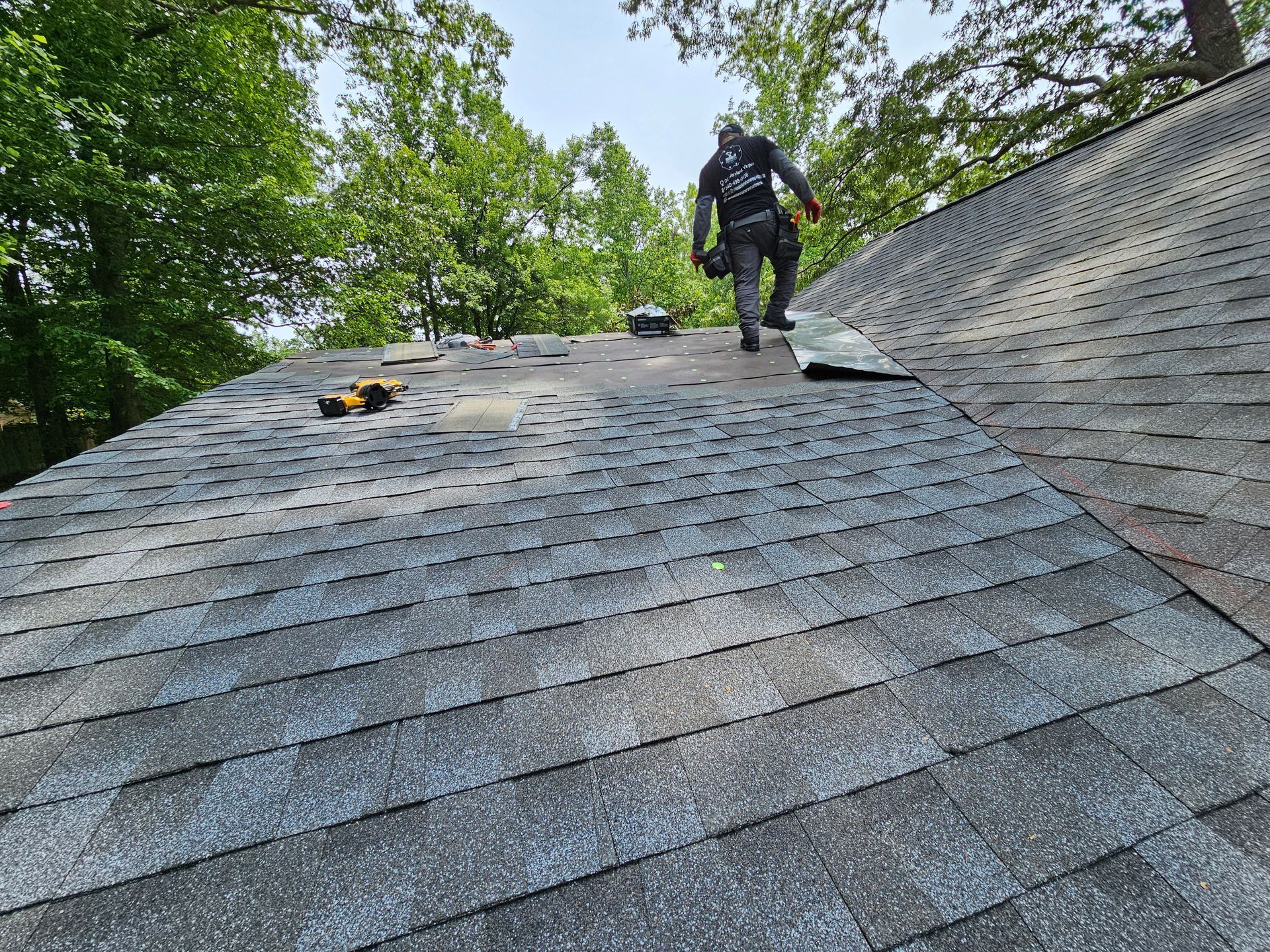 Worker on a roof beside trees, inspecting gray shingles on a steep slope.