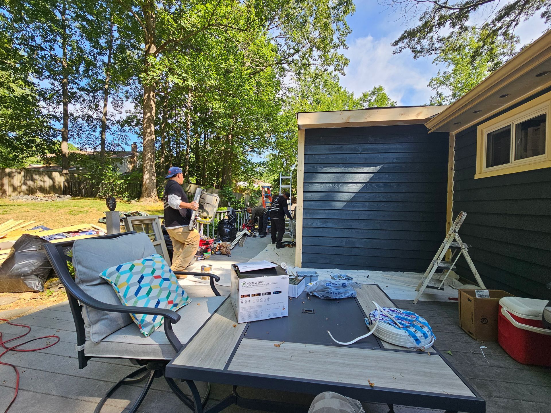 Backyard patio with folding chair, boxes, and a person unloading items beside a blue house