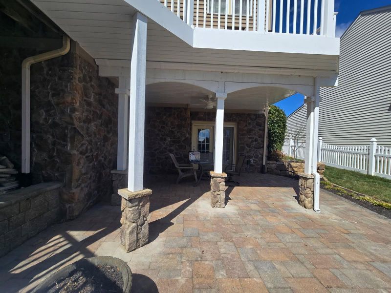Covered stone patio with white columns, brick pavers, and a staircase to an upper deck.