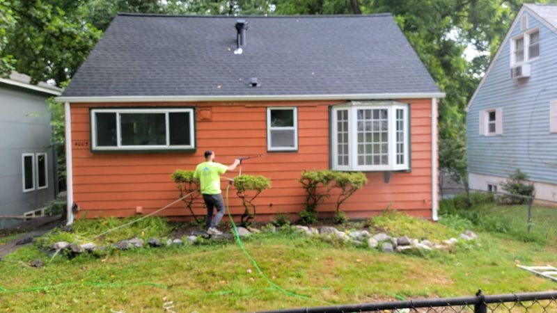 Person painting the front of an orange house with a spray gun in a leafy yard