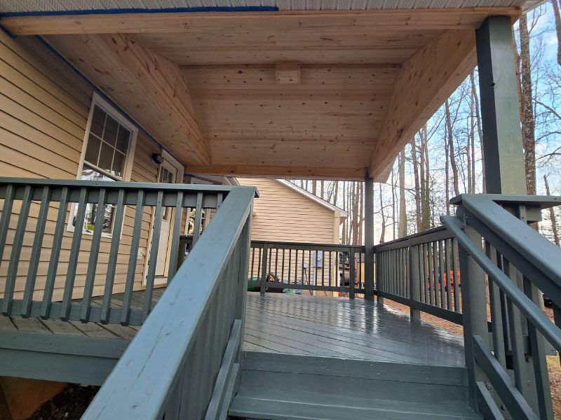 Wooden ramp and porch with railings beside a house, covered by a roof, in a wooded setting.