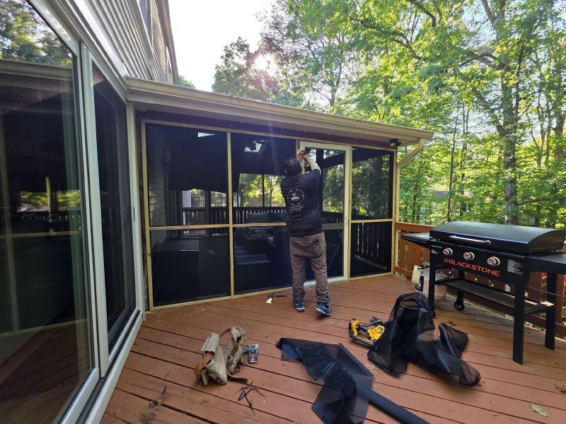 Person cleaning screened porch windows beside a grill, with tools and bags on the deck.