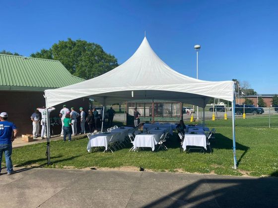 White event tent with tables set up on a grassy field, with people gathered nearby on a sunny day