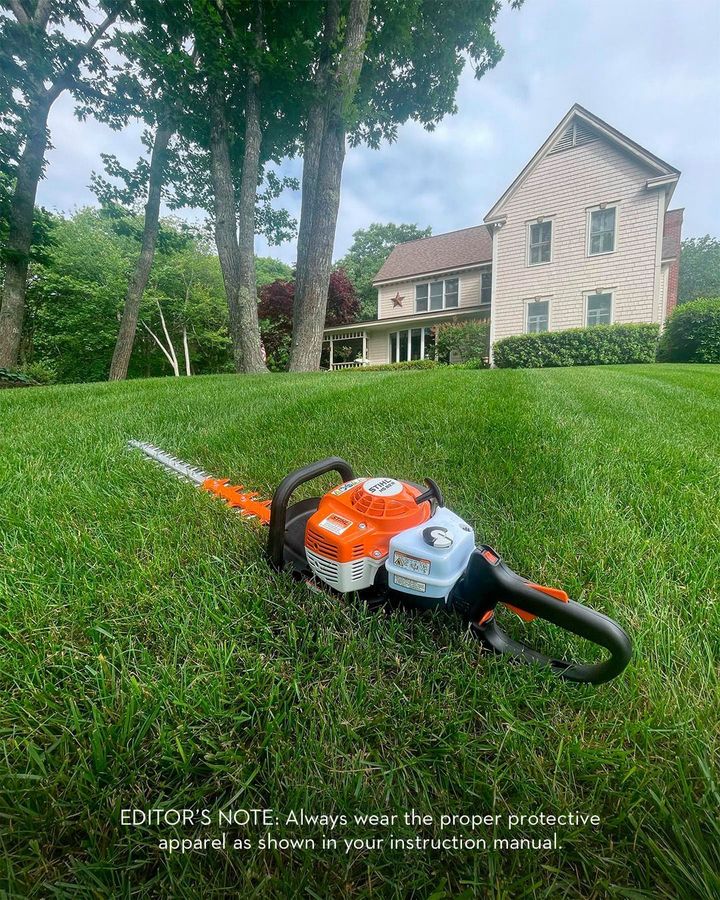 Gas-powered hedge trimmer on green lawn in front of a house and trees