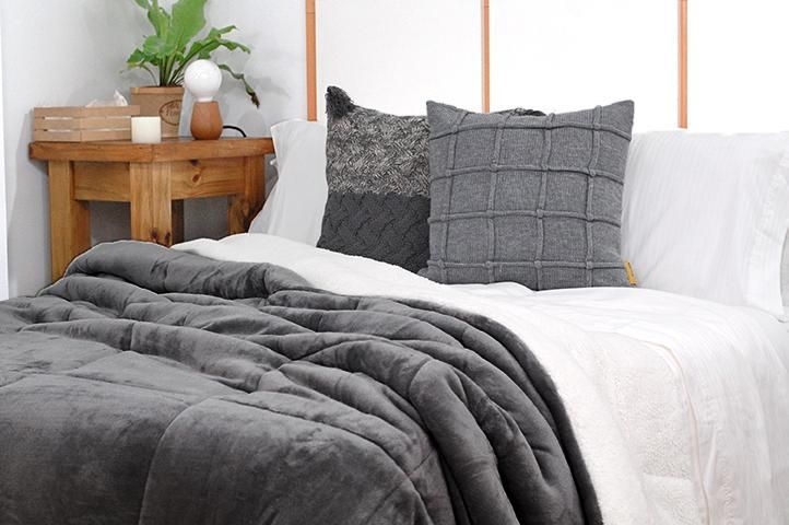 Una cama con sábanas blancas y dos almohadas grises, una manta gris y una mesita de noche de madera.