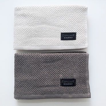 Dos paños de cocina doblados, uno color crema y otro gris, con la etiqueta