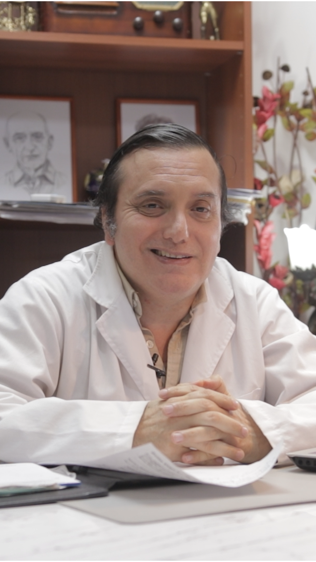 Un médico sonriente con bata blanca, sentado en un escritorio en una oficina, con estanterías y flores detrás de él.
