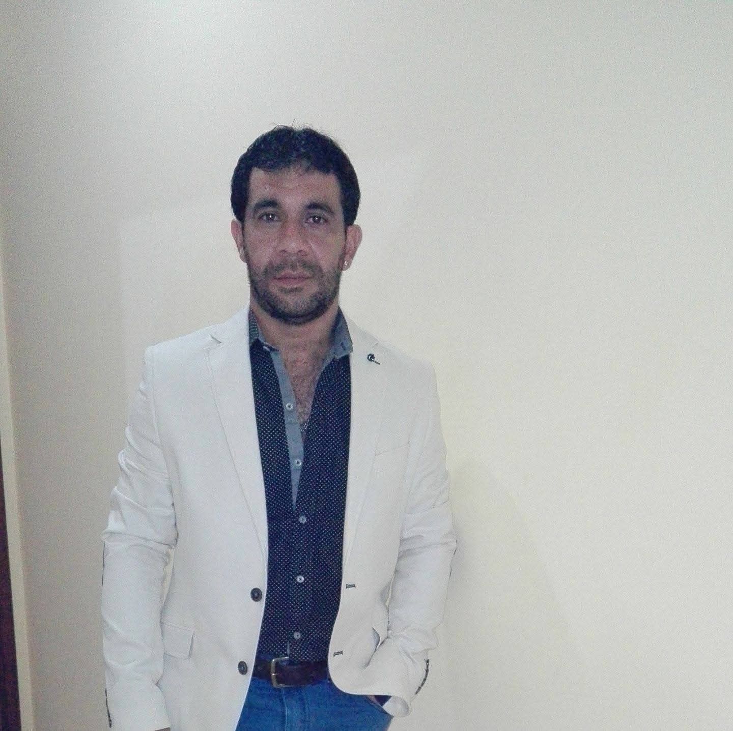 Un médico sonriente, con bata blanca y camisa rosa, sentado detrás de un libro abierto en un escritorio de una oficina.