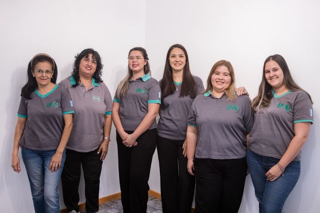Seis mujeres con polos grises y pantalones negros posan juntas, sonriendo, frente a un fondo blanco liso.