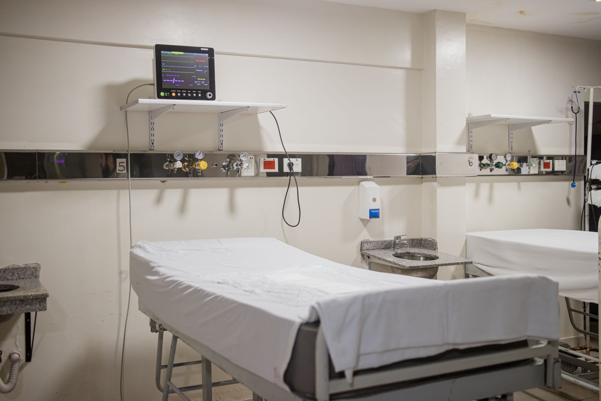 Cama de hospital con sábanas blancas y manta azul junto a una cortina de privacidad en una habitación clínica.