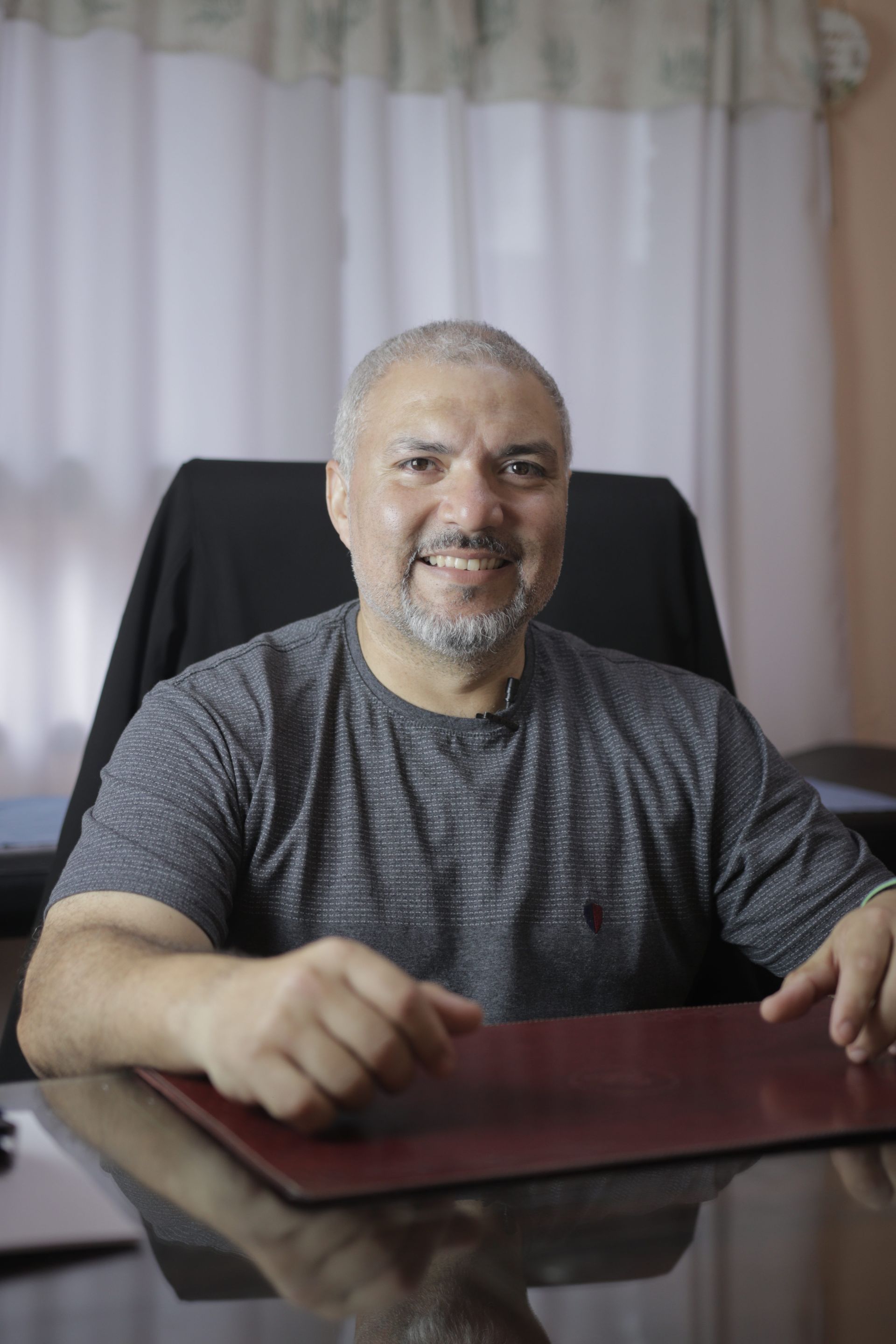 Un hombre sonriente está sentado en un escritorio con una computadora portátil, frente a unas cortinas blancas.