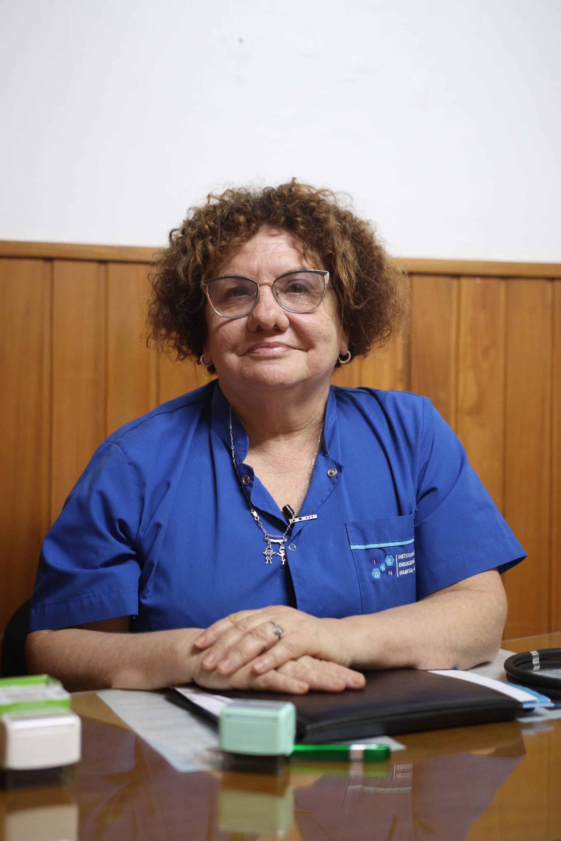 Mujer con uniforme médico azul sentada en un escritorio, mirando a la cámara con las manos cruzadas.