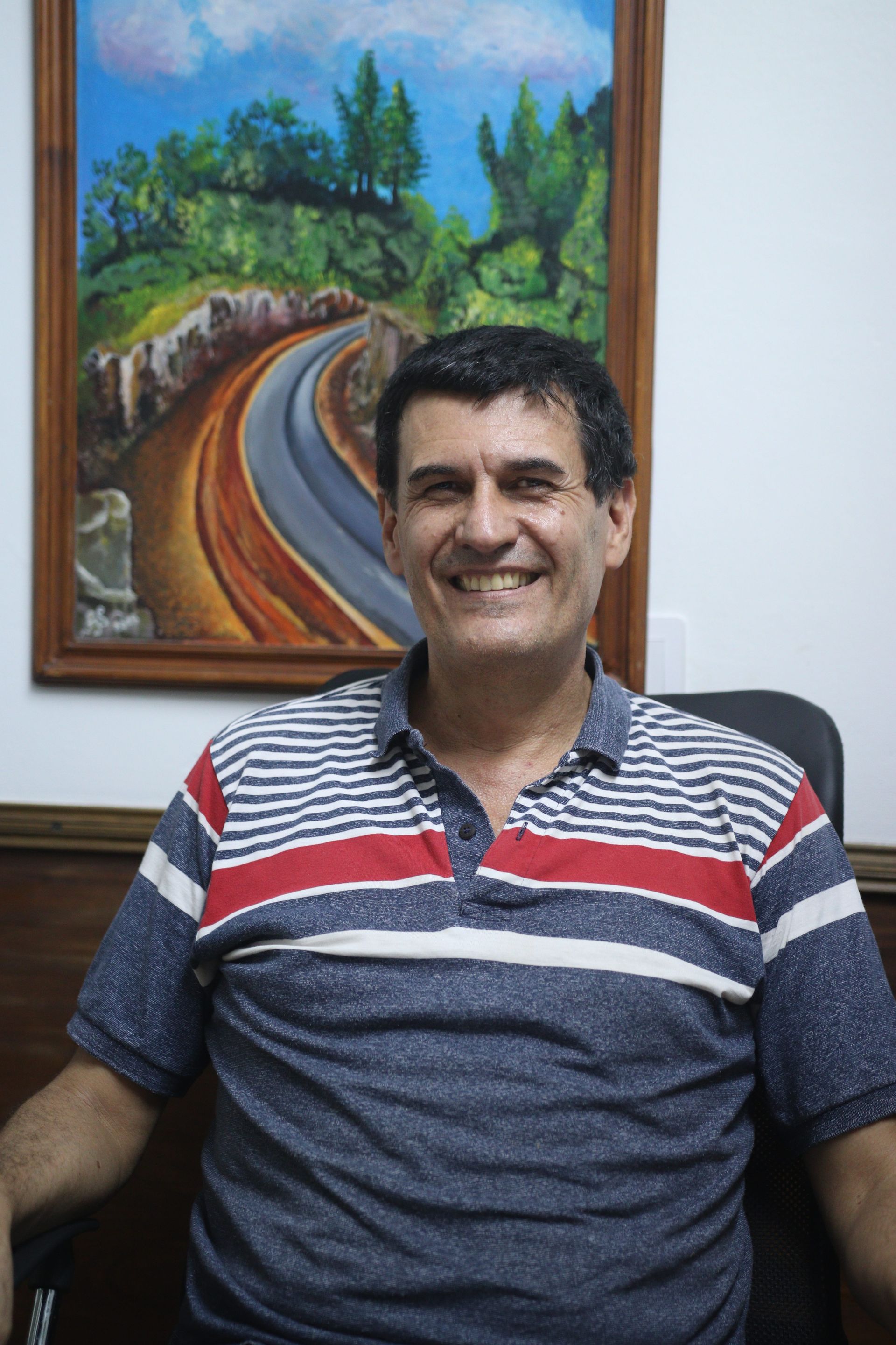 Hombre sonriente con una camisa polo a rayas sentado en el interior junto a un cuadro paisajístico enmarcado.