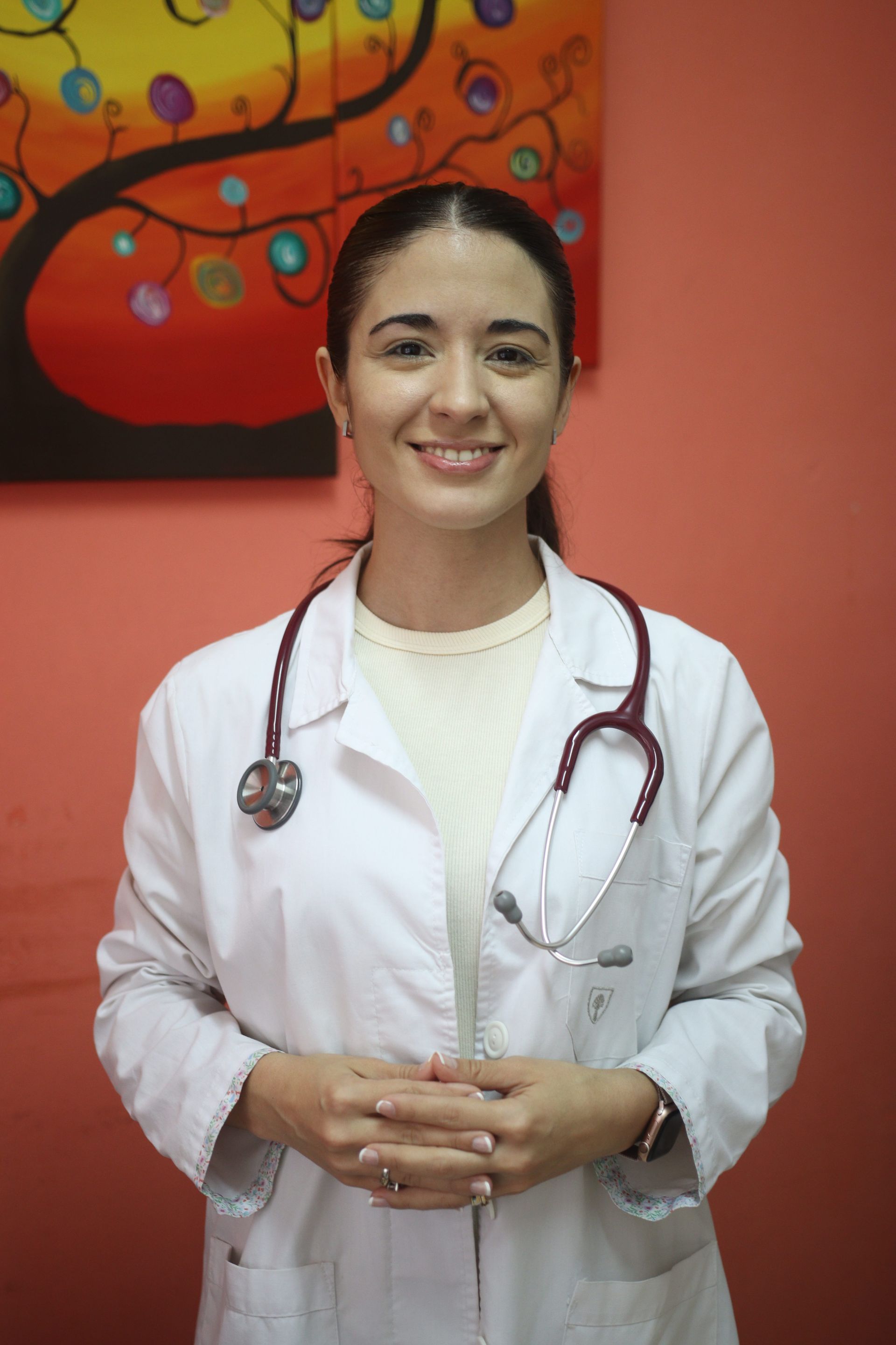 Un médico con bata blanca y estetoscopio, de pie y sonriendo contra una pared roja.