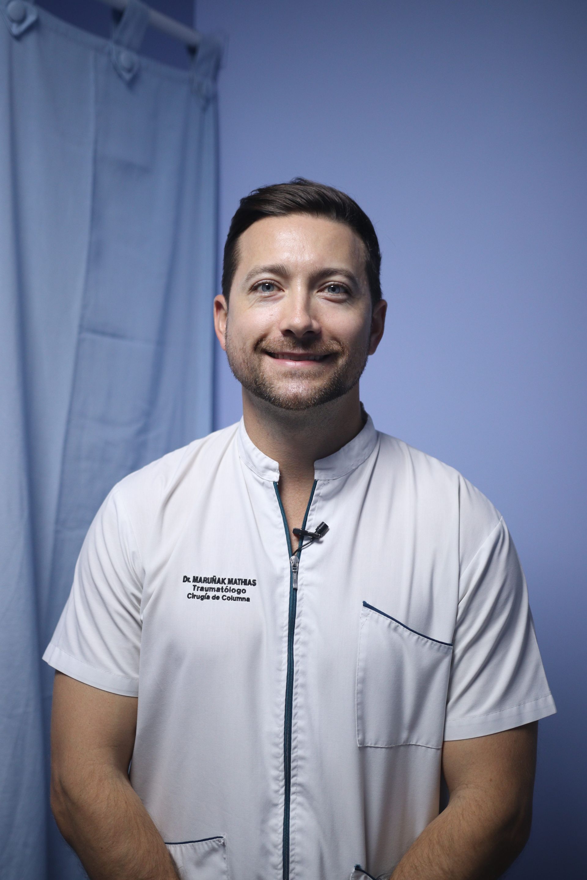 Persona vestida con uniforme médico blanco, de pie y sonriendo frente a una cortina azul.
